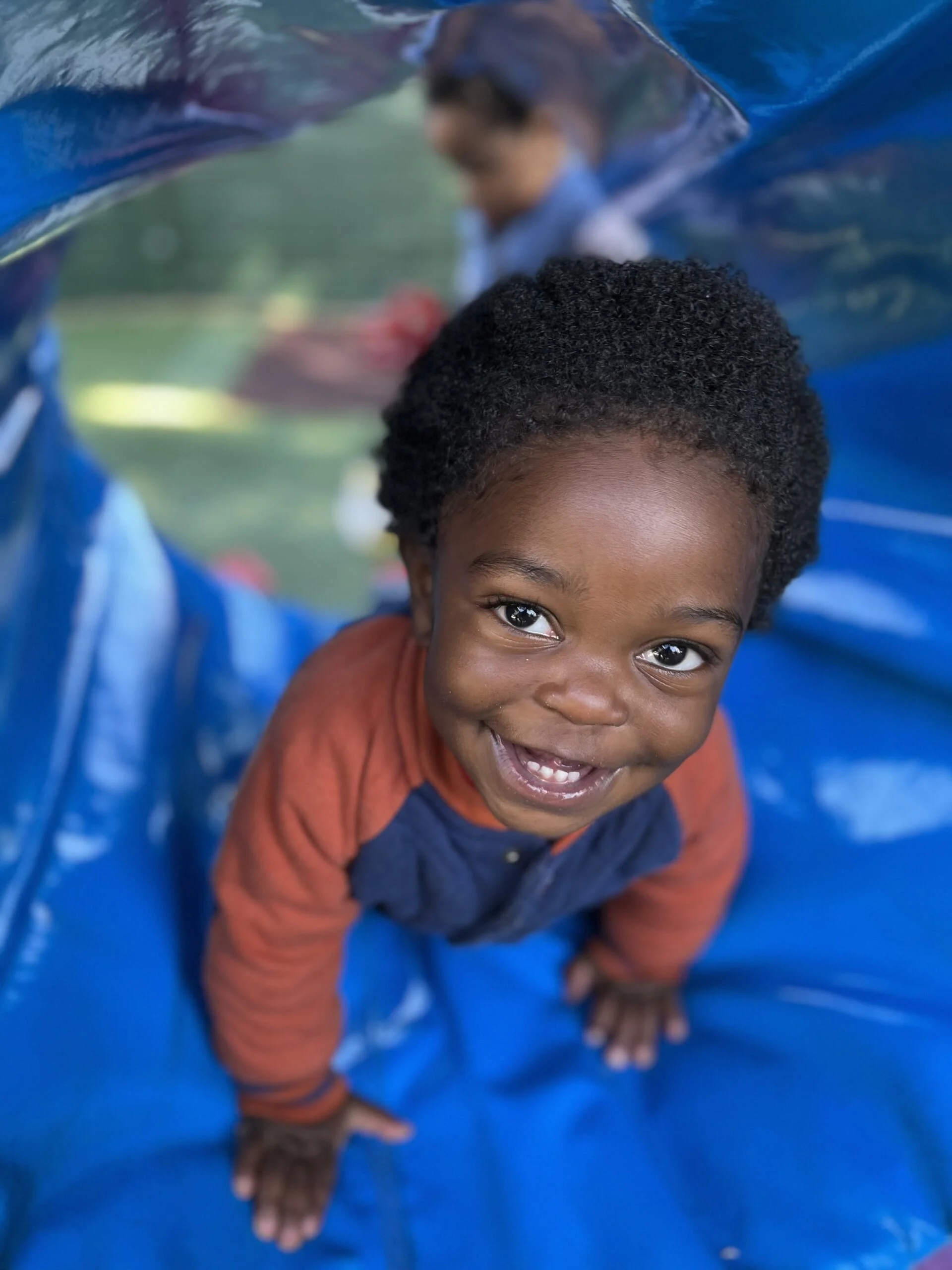 A young boy with curly black hair and a bright smile, crawling on a blue plastic tunnel or slide outdoors.