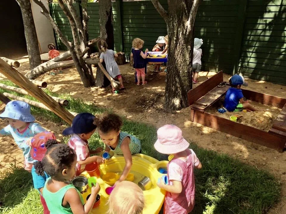 Children playing in a sand and water play area outdoors with trees and a green fence in the background.