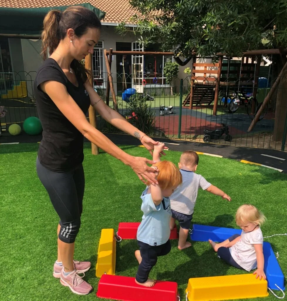 An adult woman playing with three small children outdoors on a green artificial grass playground. The woman is holding the hands of two children who are standing, while another child is sitting on the turf. There are colorful plastic obstacle bars on the ground, and playground equipment and a house are visible in the background.
