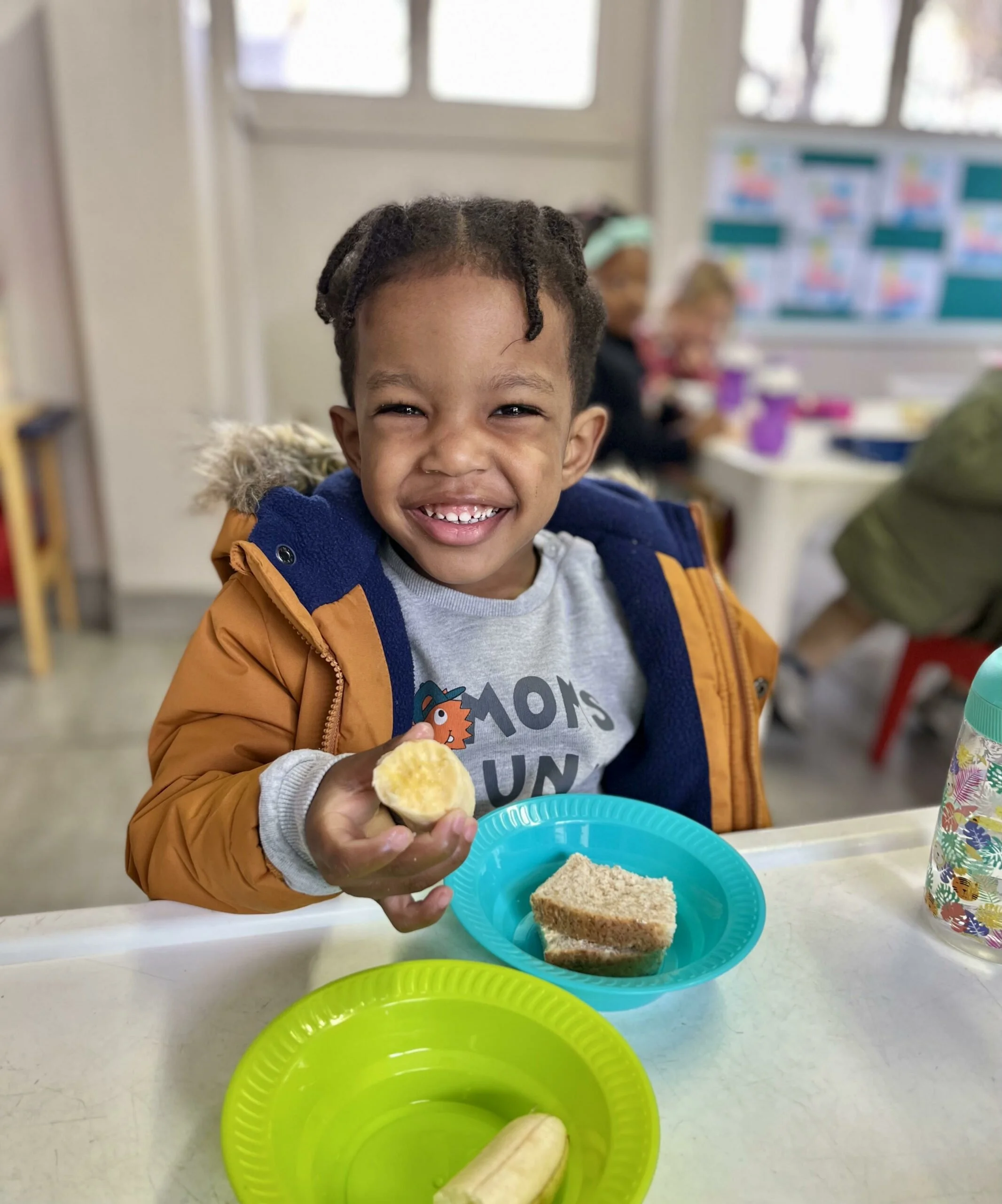 A young boy with a big smile, showing missing front teeth, sitting at a table with a slice of cake in his hand. The table has colorful plates with cake and a banana. In the background, a classroom setting with other children and a window.