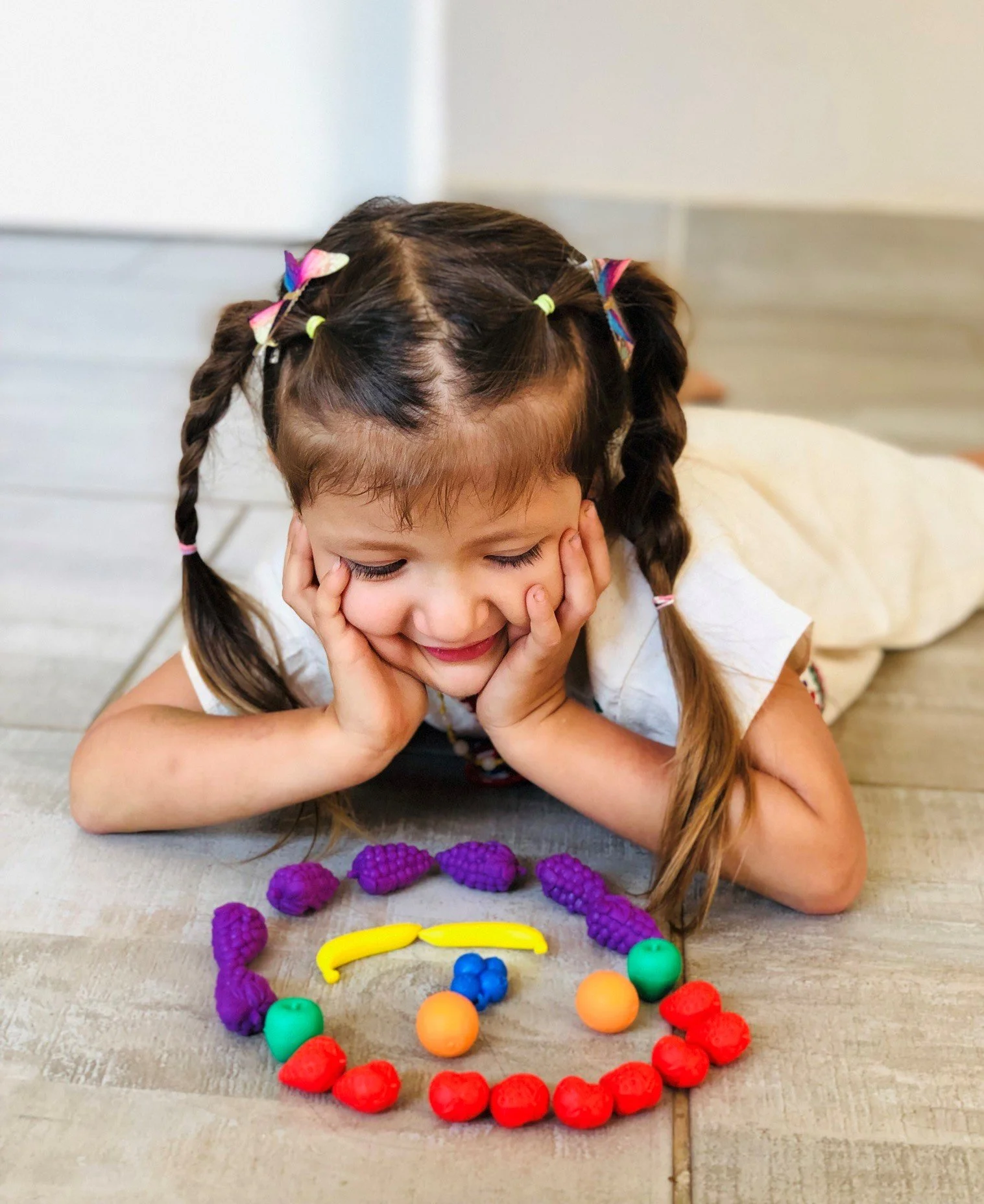 A young girl with braided hair and butterfly hair clips lying on her stomach on a wooden floor, smiling with her eyes closed, surrounded by colorful textured toys arranged in the shape of a rainbow.