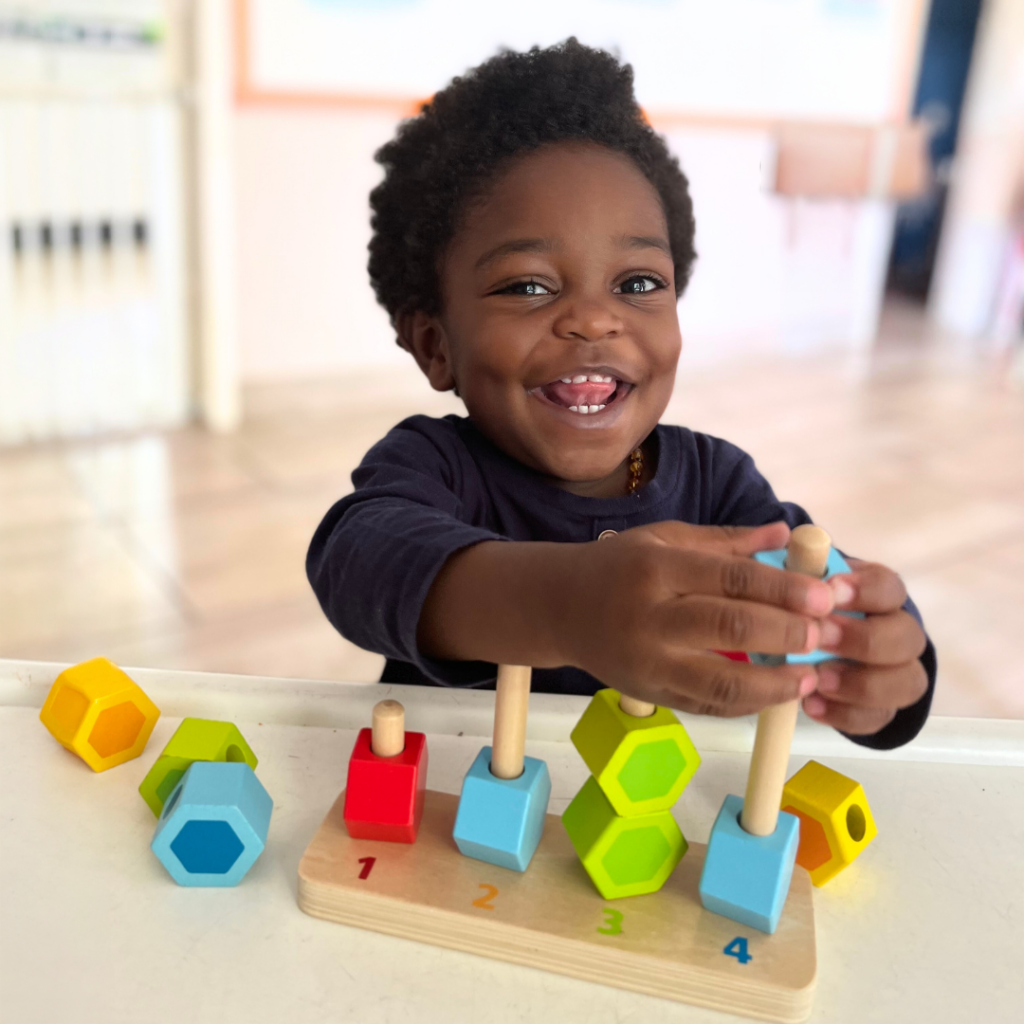 A young child with short, curly hair is smiling while playing with colorful educational toys on a table. The toys include painted wooden blocks and shaped objects in bright colors, arranged in a sequence on a wooden base with numbered steps.