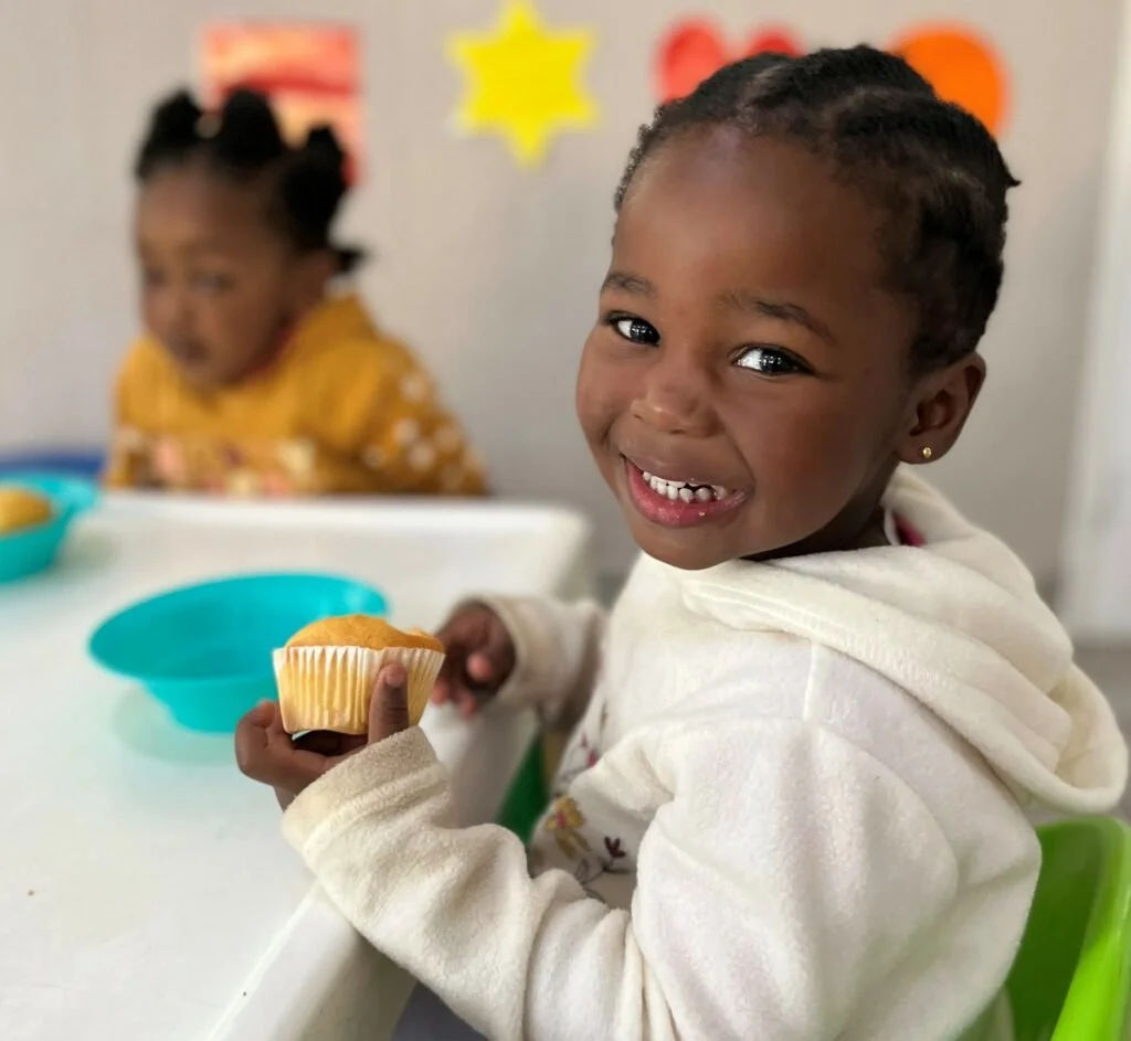 Smiling young girl with braided hair holding a cupcake at a table with teal bowls, with a blurred girl in a yellow shirt in the background.