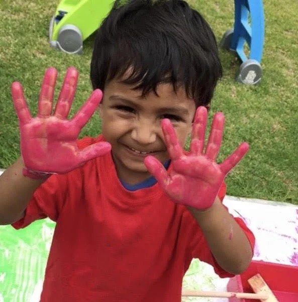 Smiling young boy with messy black hair and red shirt showing his hands covered in pink paint.