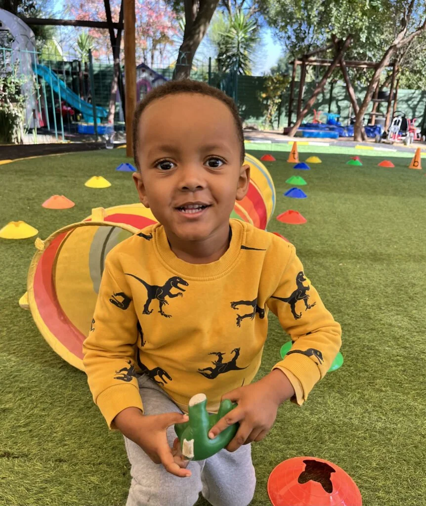 A young boy playing outside on a grassy area with colorful cones and playground equipment in the background, holding a green plastic toy.