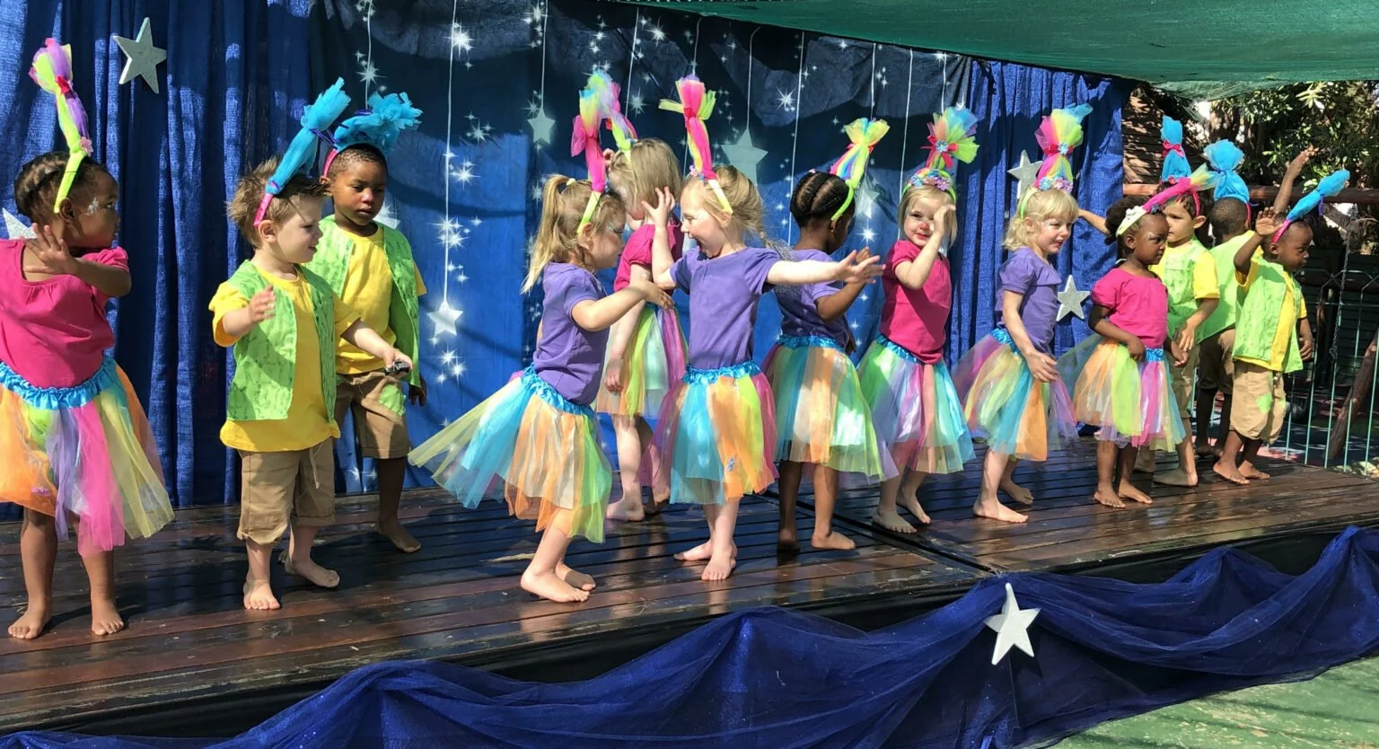 Children performing a dance or play on a decorated stage with a starry space theme, wearing colorful costumes with tulle skirts, headbands, and shirts, some are dancing and others are standing or talking.