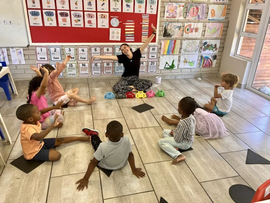 A teacher sitting on the floor in a classroom with six young children sitting on the floor around her. The children are raising their hands. The classroom has educational posters on the wall and colorful artwork.