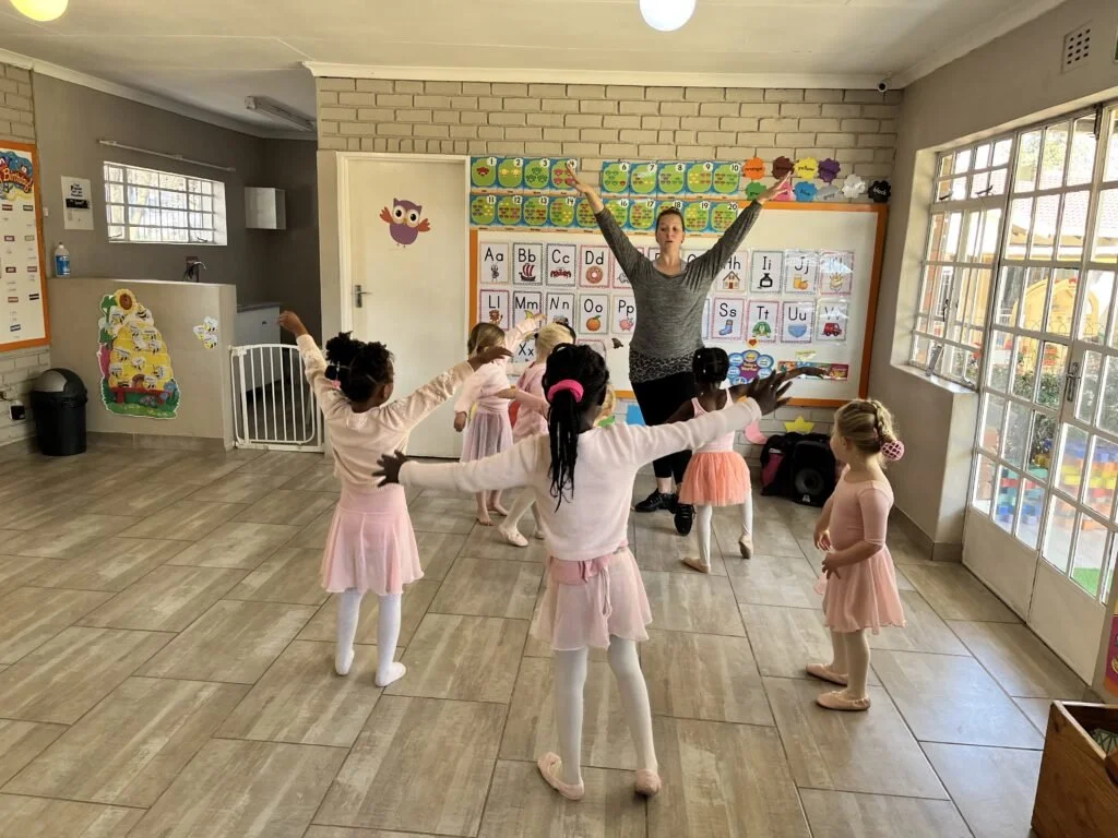 A dance or ballet class for young children with an instructor leading the children in a pose, inside a classroom decorated with educational posters and a whiteboard.