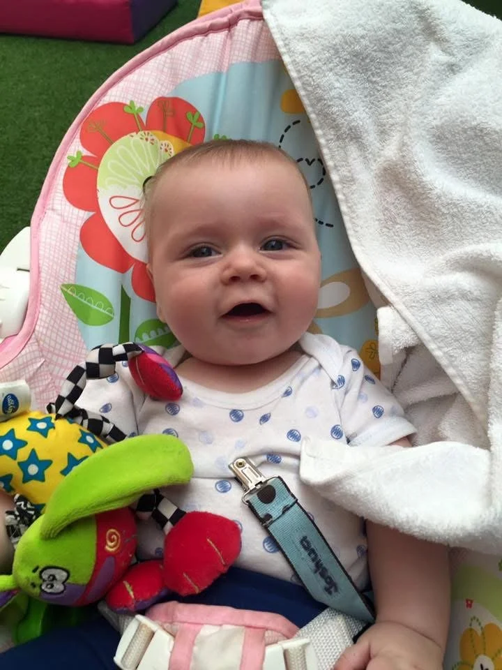 Smiling baby with light skin and short hair lying in a colorful baby seat, surrounded by plush toys, with a white towel partially covering the seat.
