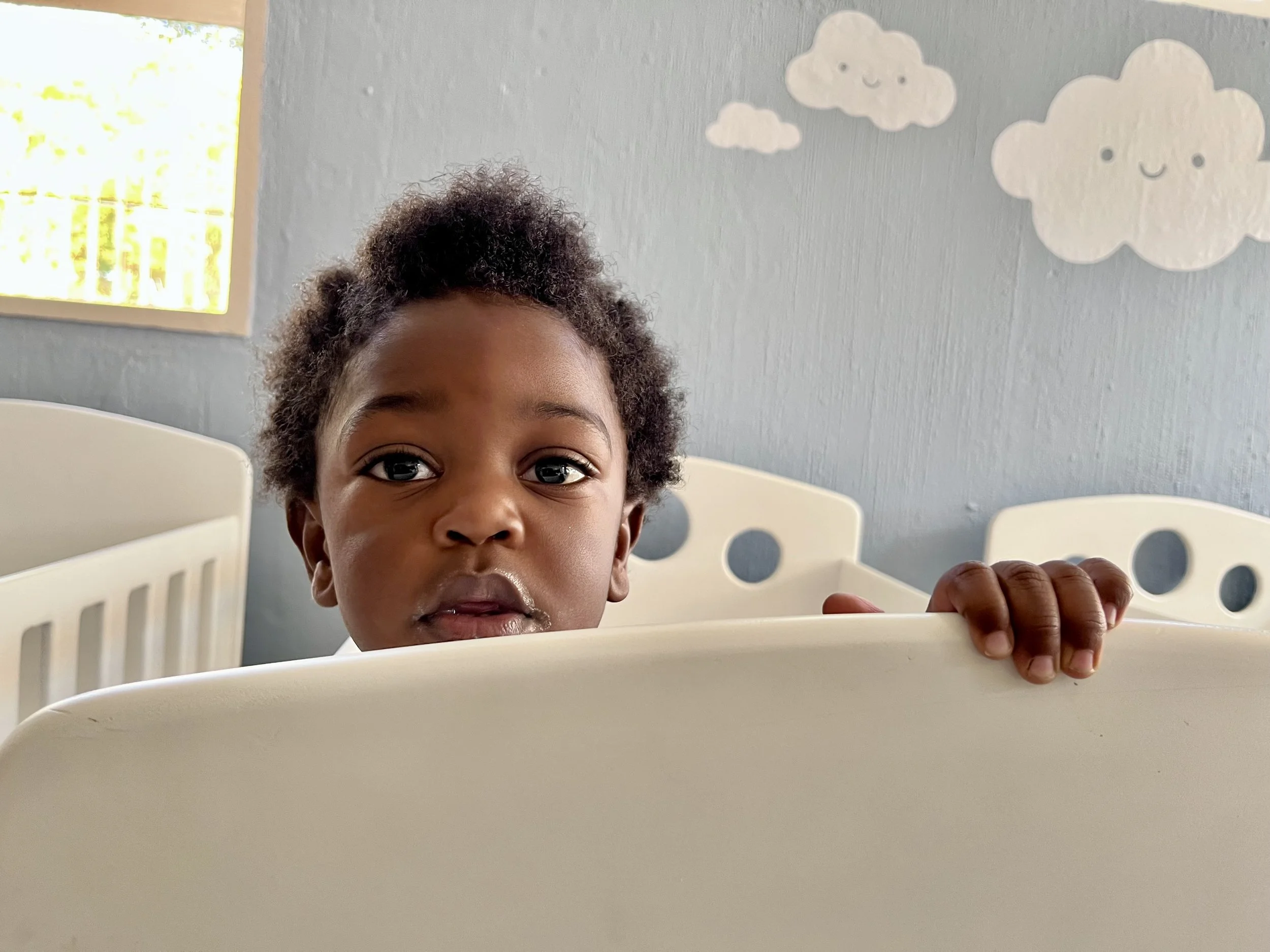 A young child with curly hair peeking over the top of a white plastic object, possibly a container or a guardrail, in a room with light blue walls decorated with paper clouds featuring smiling faces.