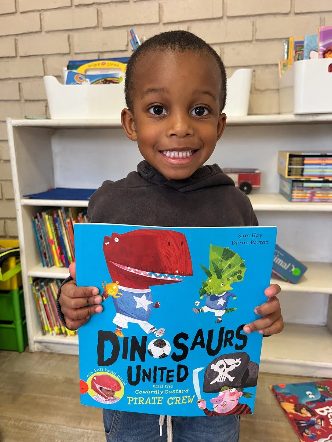 A smiling young boy holding a children’s book titled "Dinosaurs United and the Cowardly Custard Pirate Crew." The background shows a bookshelf with other books and toys.