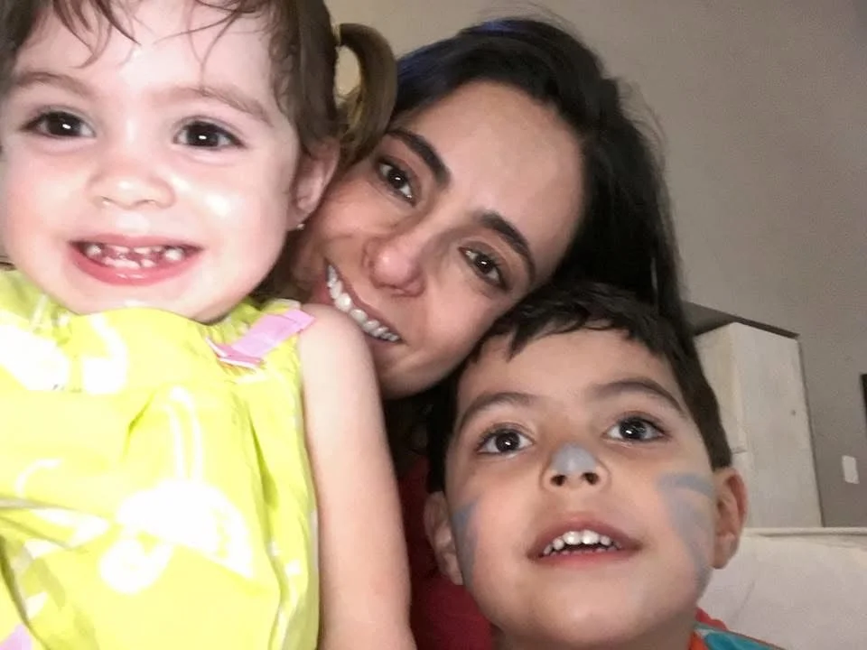 A woman with dark hair smiling and hugging two young children, one girl with curly hair and a yellow shirt, and one boy with face paint around his nose and cheeks.