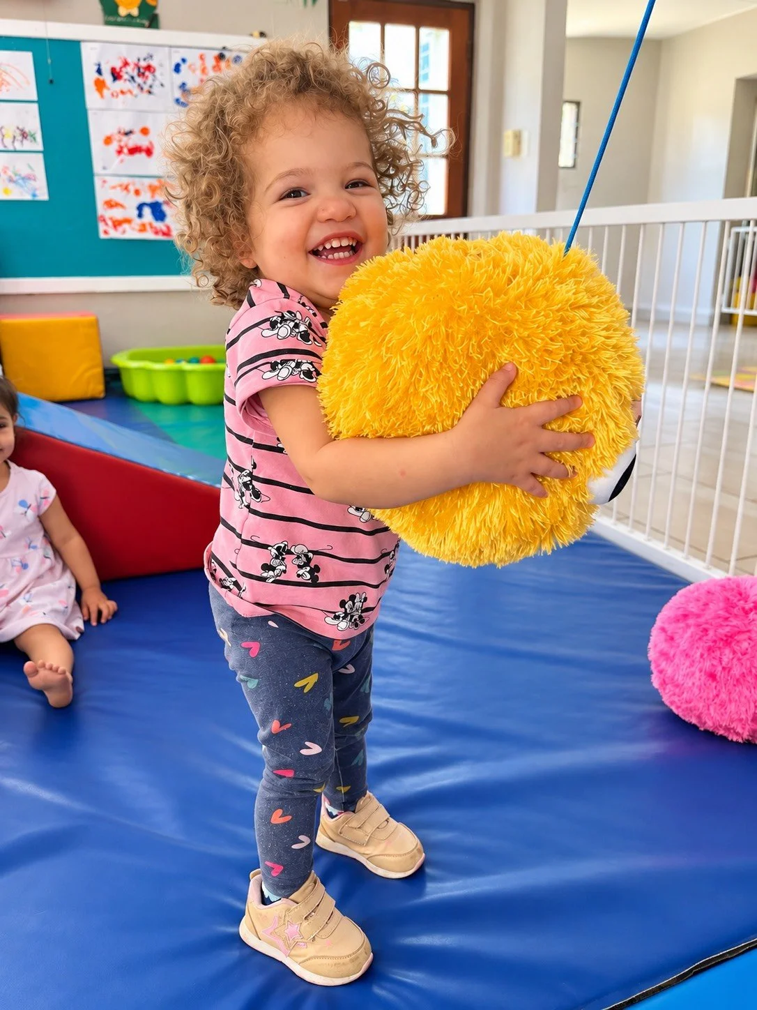 A young girl with curly hair holding a large yellow fluffy ball at an indoor play area, smiling brightly. Another child is sitting on a padded surface in the background.