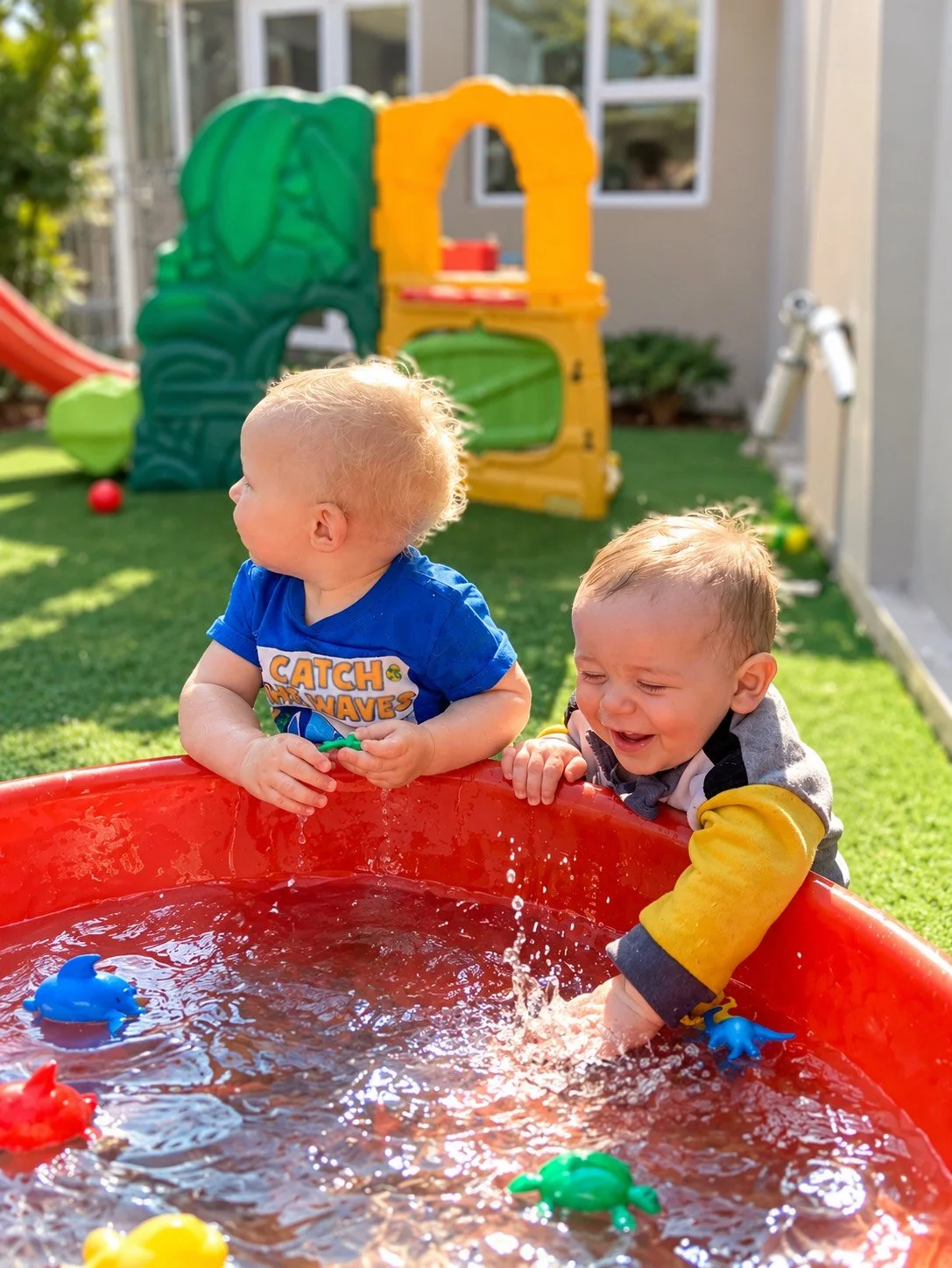 Two young children playing in a red plastic water table outdoors surrounded by toys and a green play structure, with sunlight and a house wall in the background.