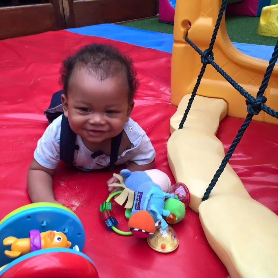 A baby crawling on a colorful play mat outside, surrounded by baby toys and play equipment.