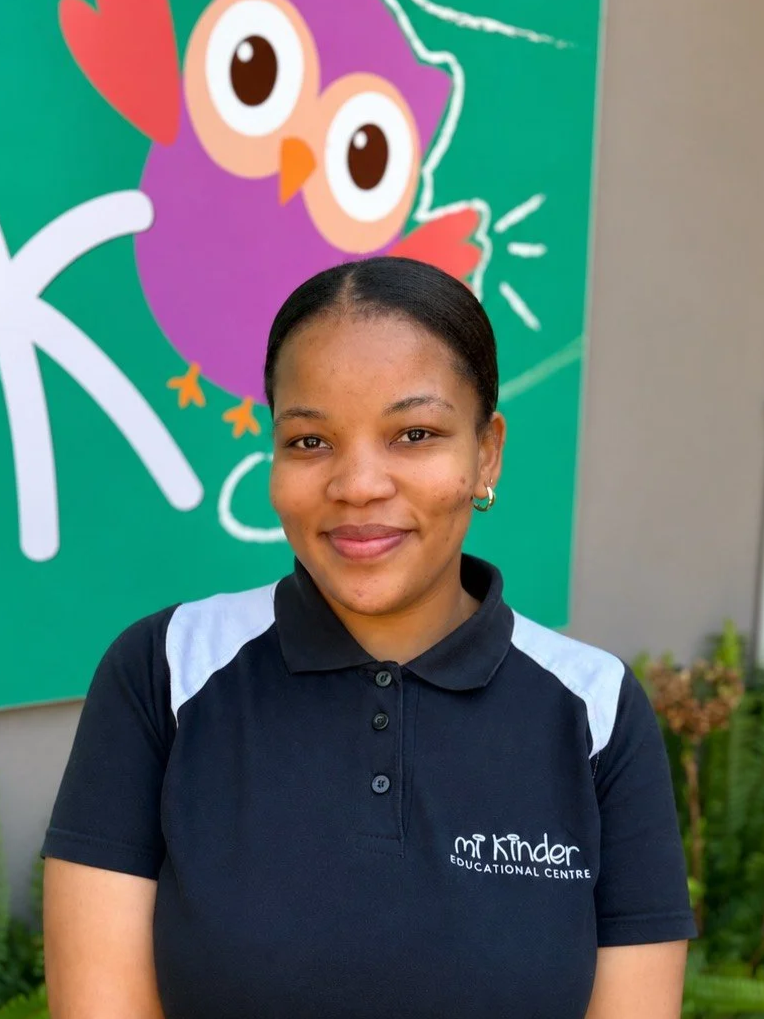 A young woman with dark hair tied back, wearing a navy blue polo shirt with white accents, standing outdoors in front of a colorful cartoon owl illustration on a green background. The shirt has the logo 'Mt Kinder Educational Centre'.