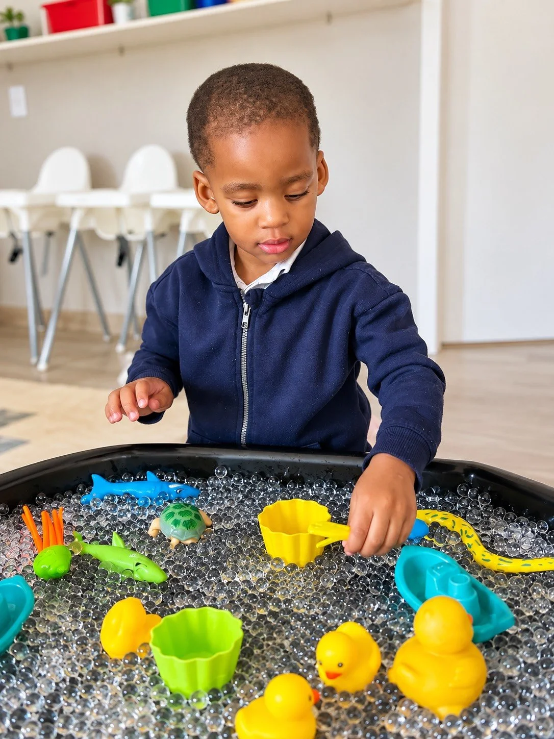 Young boy playing with floating rubber ducks, plastic sea animals, and scoops in a sensory water table.