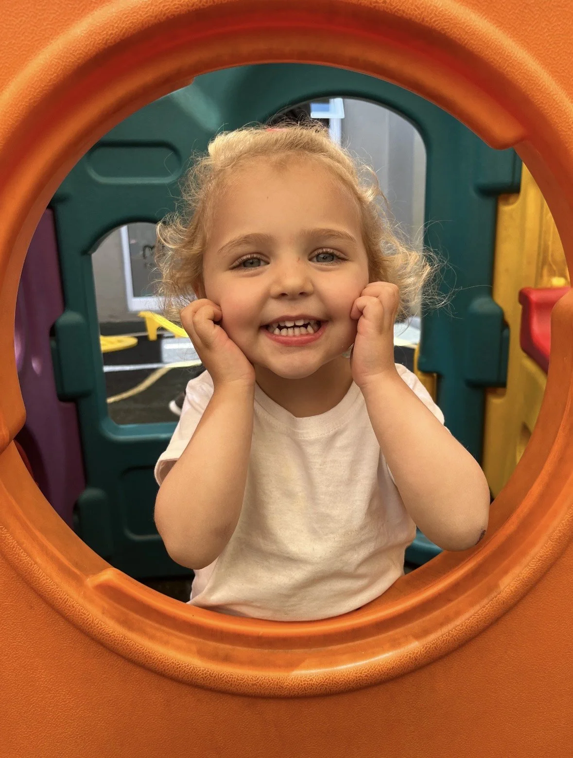 A young girl with curly blonde hair smiling and holding her face with her hands, sitting inside a colorful playground structure.