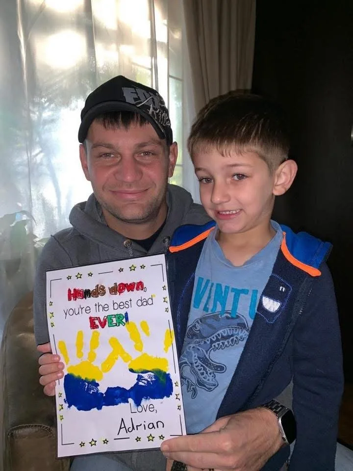 A man and a boy holding a handmade card. The card has colorful writing that says, 'Hands down you're the best dad EVER! Love, Adrian', with yellow handprints at the bottom.