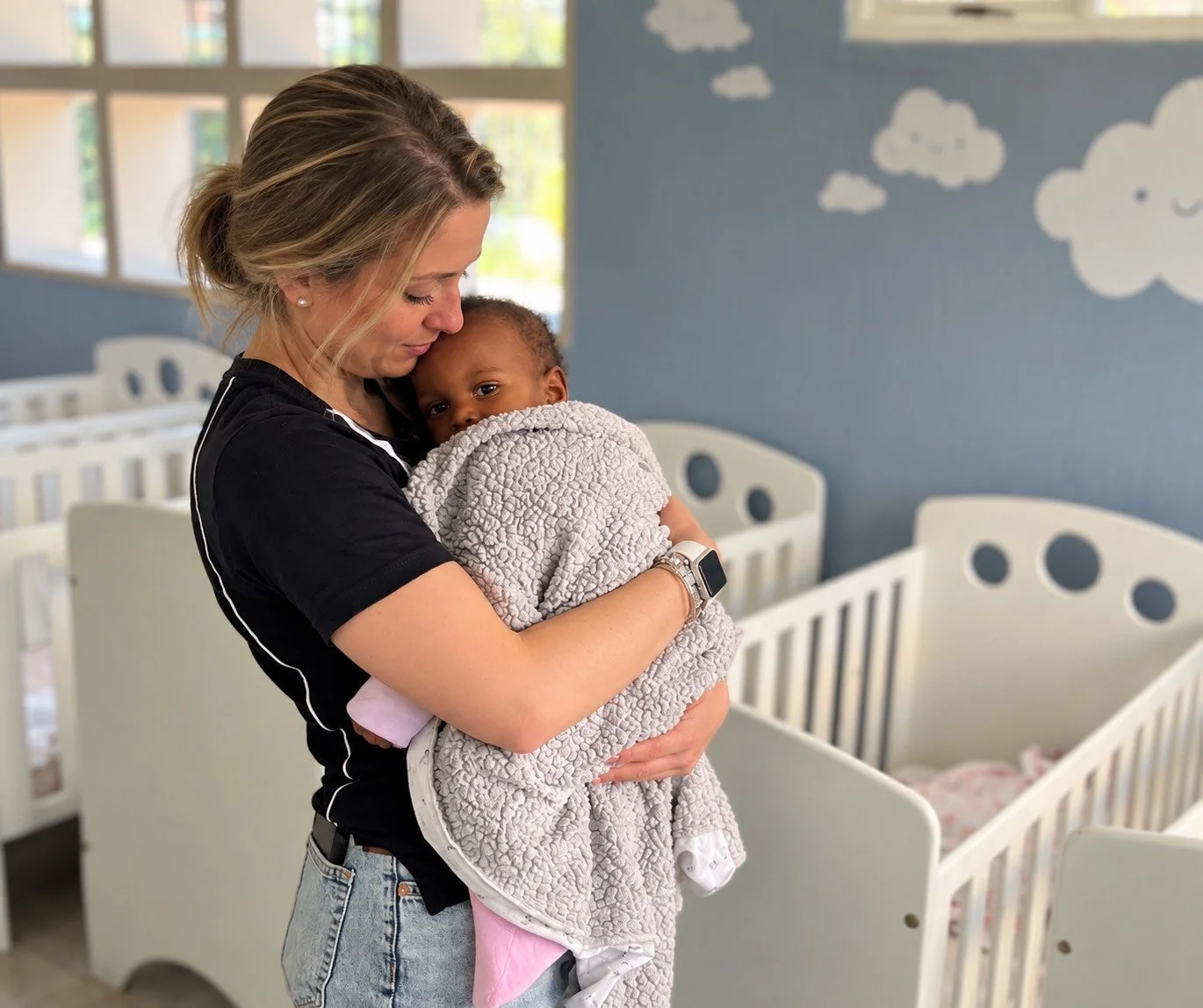A woman holding and comforting a young child in a nursery or daycare room, with cribs and cloud-themed wall decorations in the background.