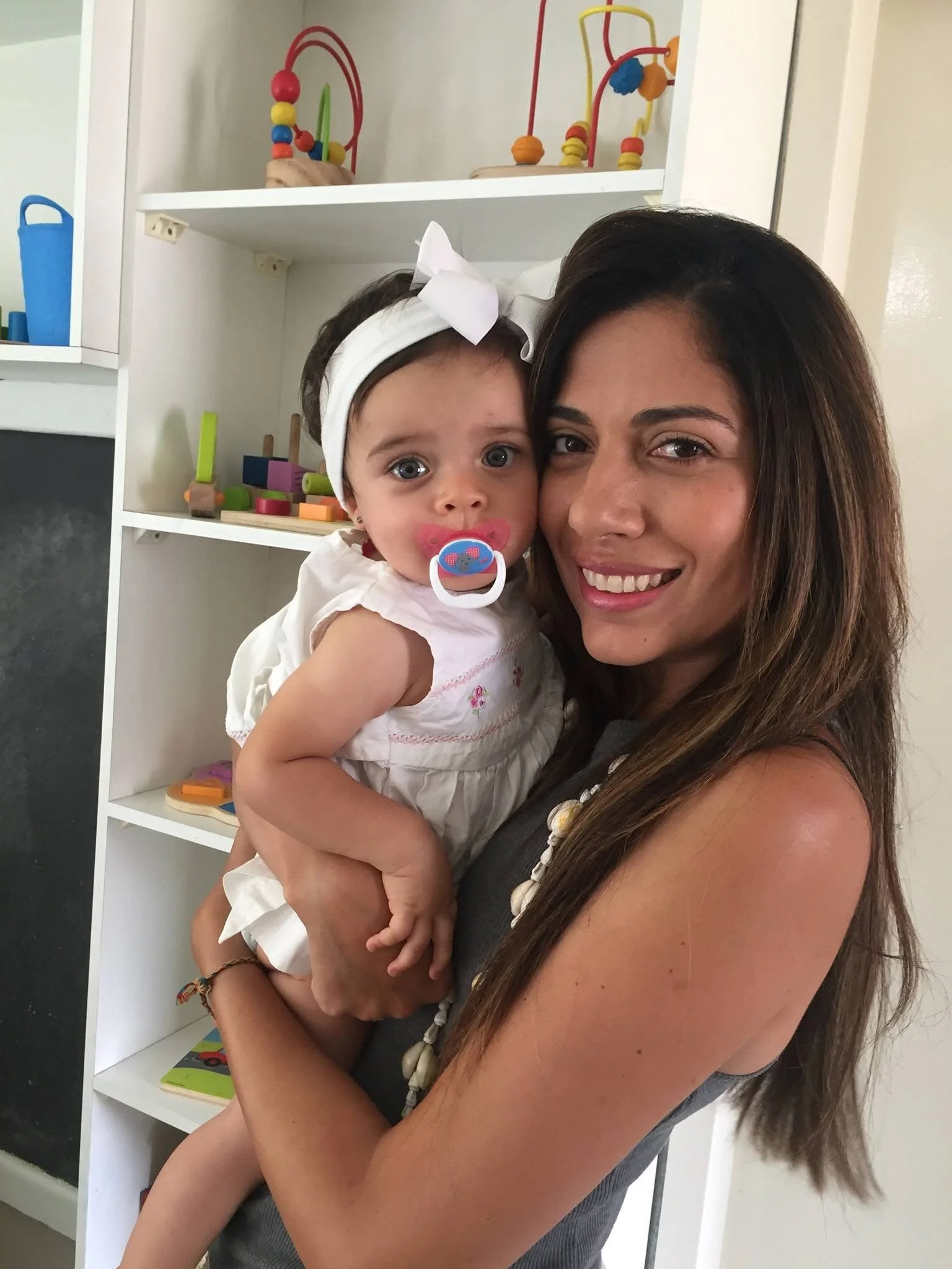 A woman holding a young girl with a pacifier in her mouth, both smiling, in front of white shelves filled with colorful toys.