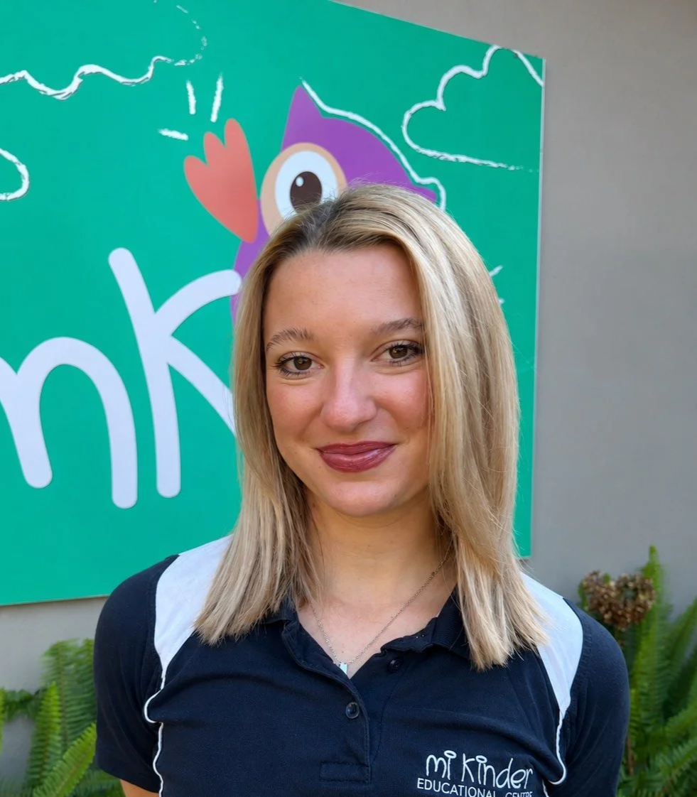 A woman with blonde hair and a dark polo shirt standing outdoors in front of a colorful sign with a cartoon owl and the words 'Mi Kinder Education Center'.