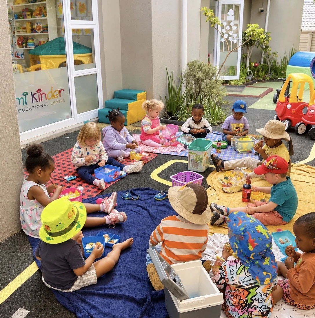 Children sitting on blankets and mats outdoors, having a snack or lunch during a school break at a childcare center named 'Mini Kinder Educational Centre.'
