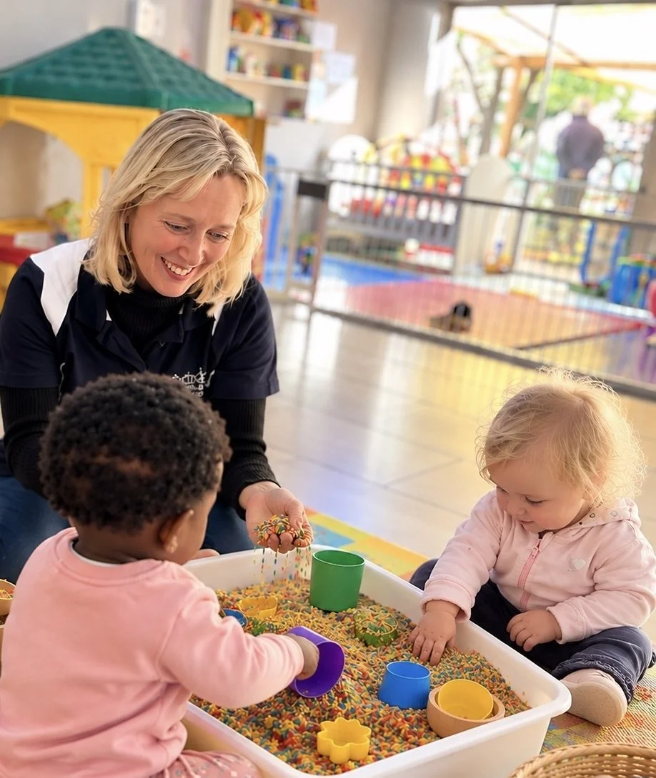 A woman playing with two young children at a sensory table filled with colorful rice or beans, in a play area with a playground in the background.