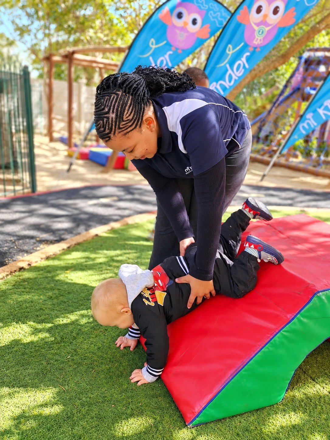 A woman is helping a toddler onto a red and green padded climbing wedge outdoors on a grassy area during the daytime. There are colorful flags with owl designs and a playground structure in the background.