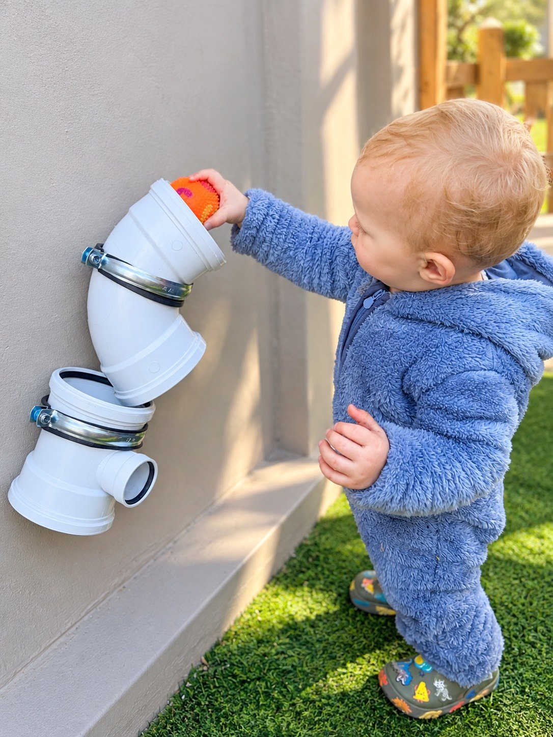 A young child in a blue fuzzy coat and colorful rain boots is placing a small orange ball into a brown pipe connected to a wall, which is part of a makeshift outdoor pipe system.