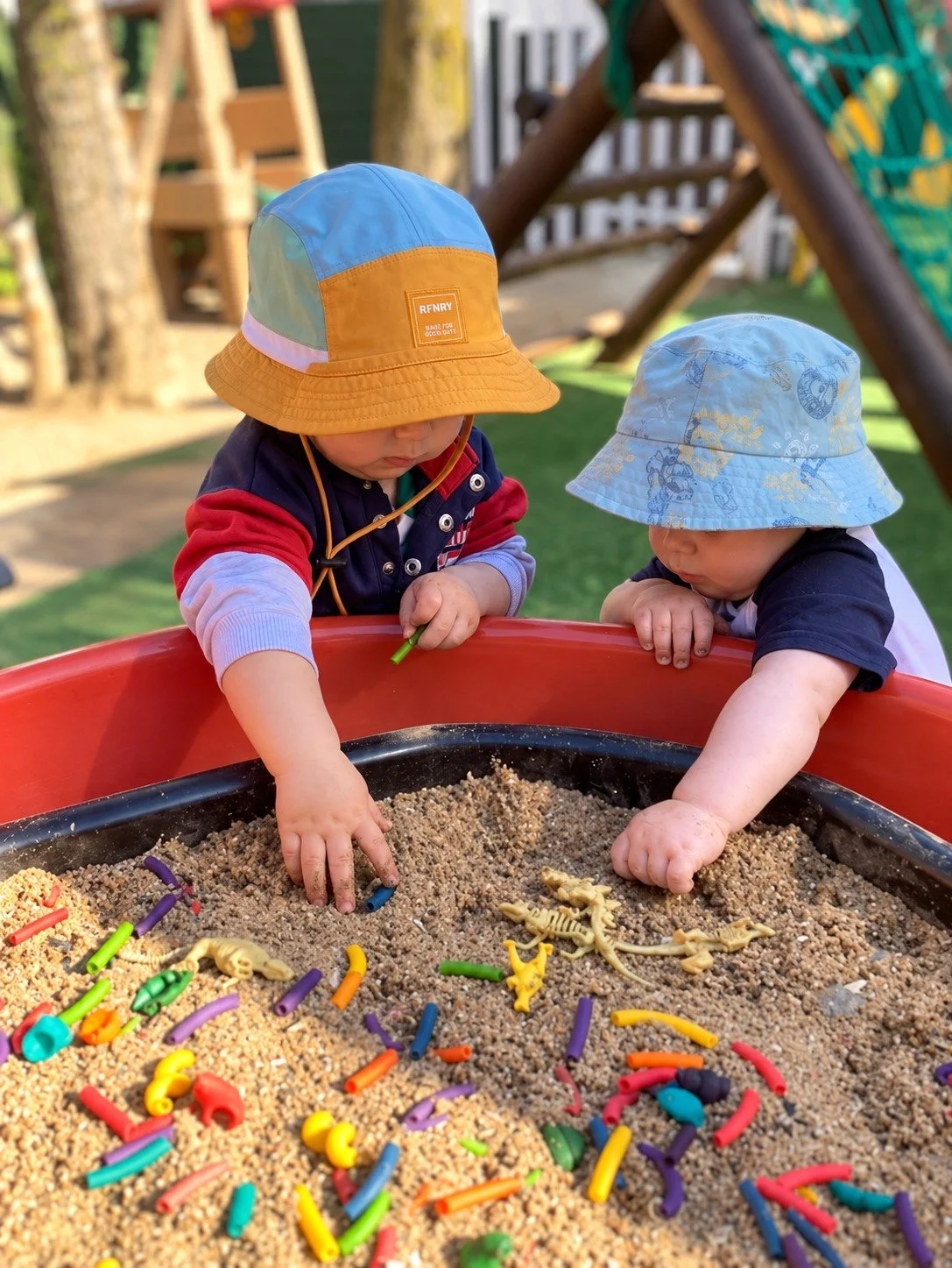 Two young children playing in a sandbox filled with colorful plastic toys, wearing sun hats, outdoors in a backyard with play structure and trees.