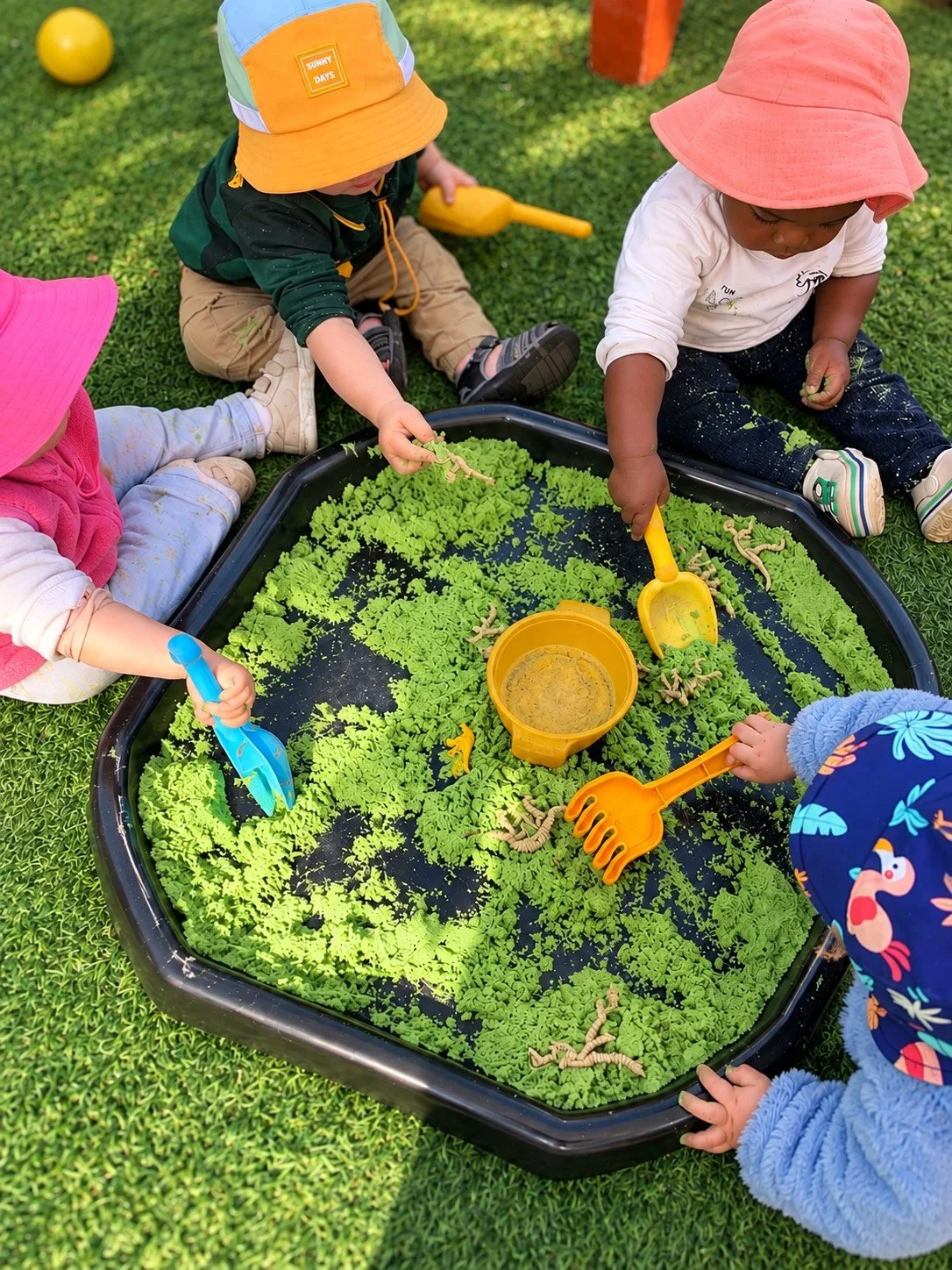 Group of young children playing with toy worms and digging in a tray filled with green plastic leaves and sand outdoors, on a grassy surface.