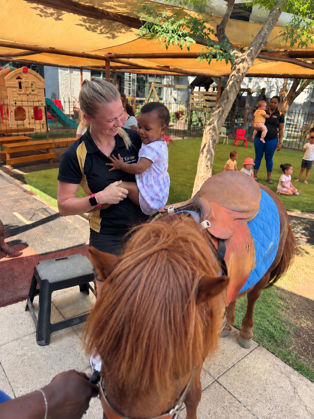 A young girl being lifted by a woman in a black and gold uniform at a children’s outdoor play area, with a pony in the foreground and other children and caregivers in the background.
