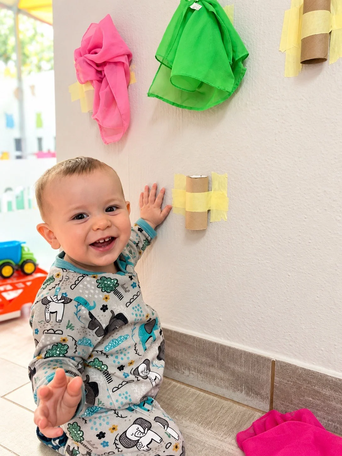 A smiling toddler in patterned pajamas sitting on a tiled floor, touching a decorated wall with painted tissue paper flowers and taped paper tubes, with toys in the background.