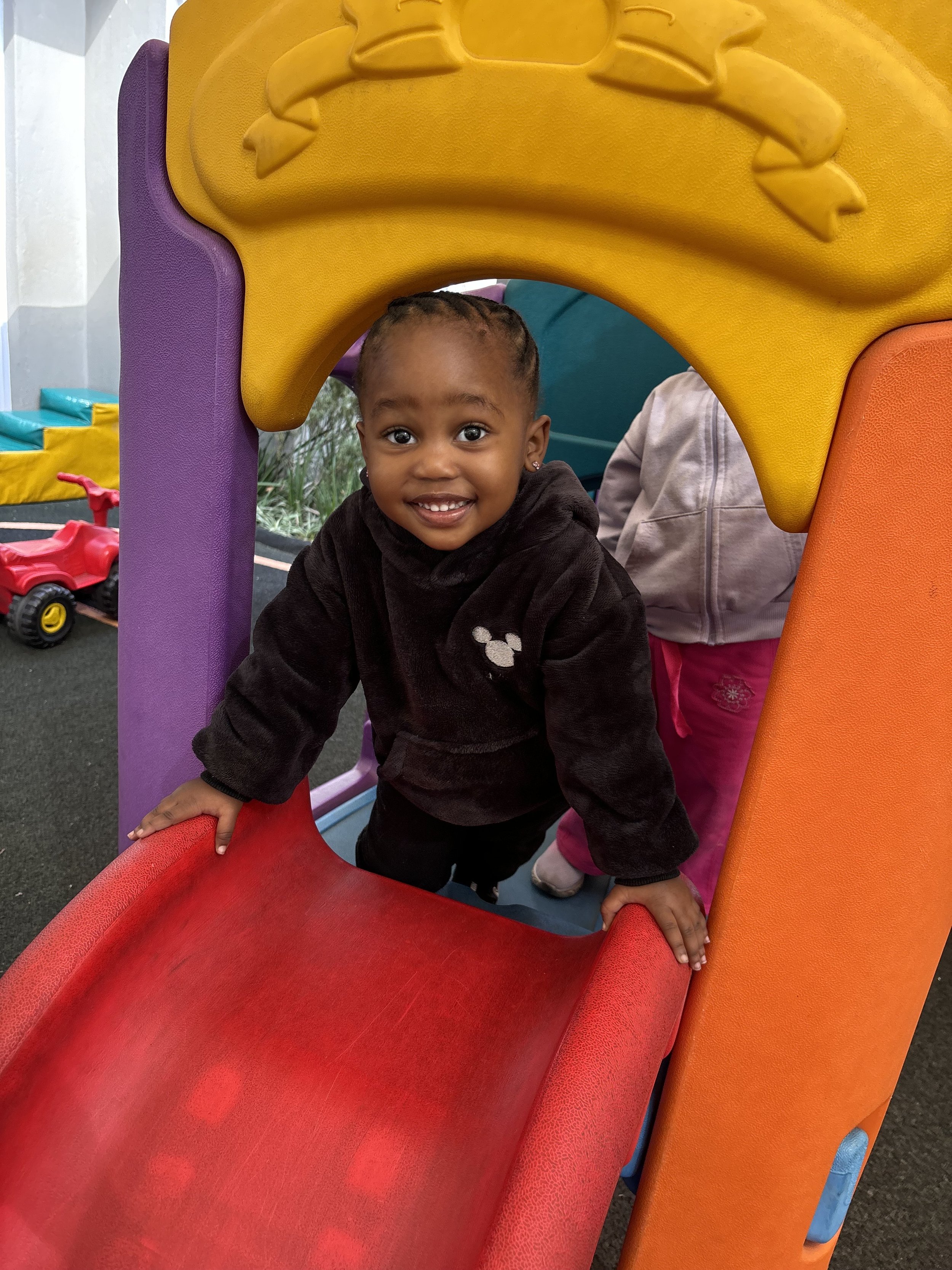 Young girl with braided hair and earrings smiling, playing on a colorful playground slide with purple, yellow, orange, and red parts.