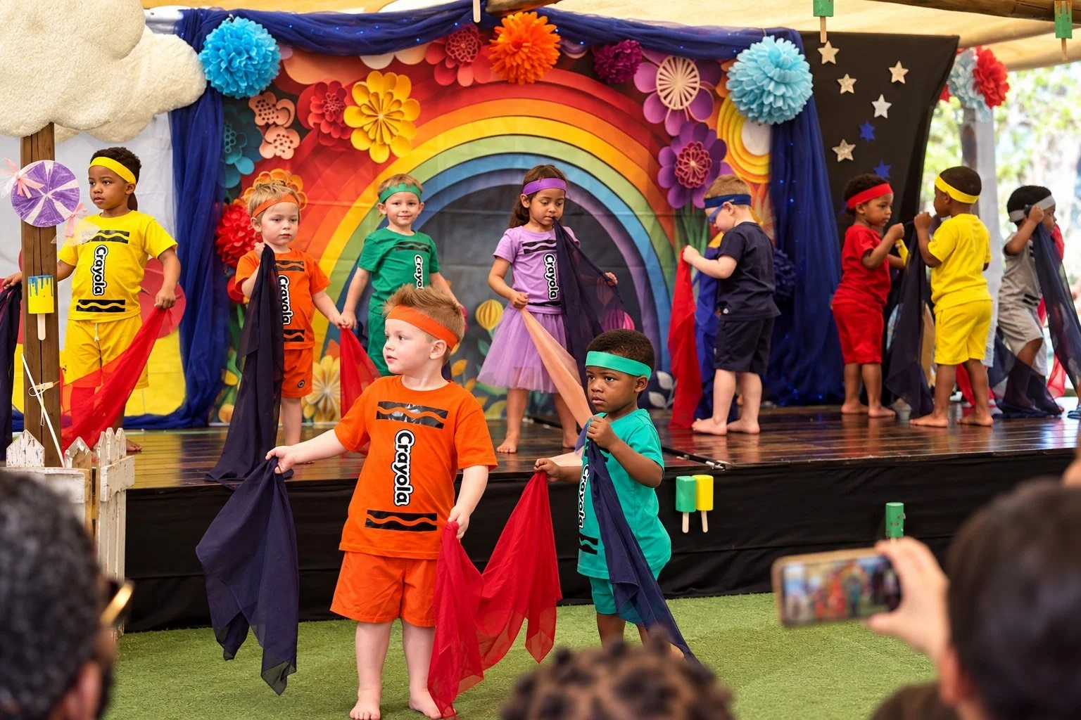 Children dressed in colorful clothing performing a stage play or dance on outdoor stage with rainbow and floral decorations, with some holding flags and wearing headbands.