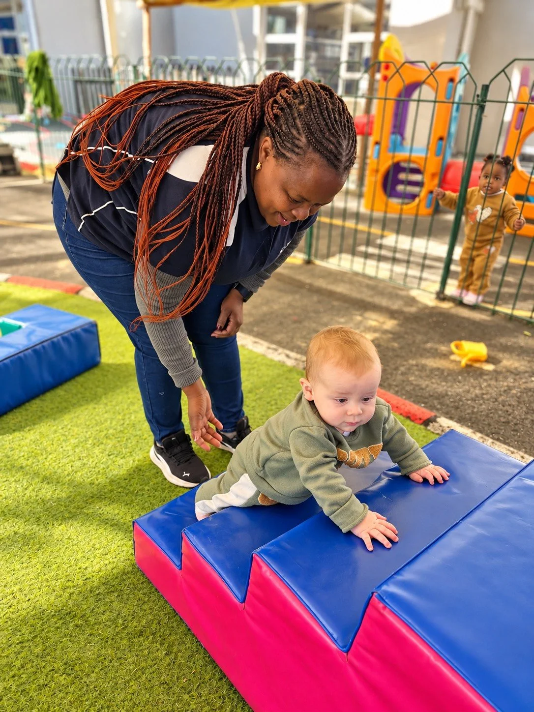 A woman helping a toddler climb onto a blue and pink padded play structure outside at a daycare or playground. In the background, a small child is peering through a fence near a colorful playset.