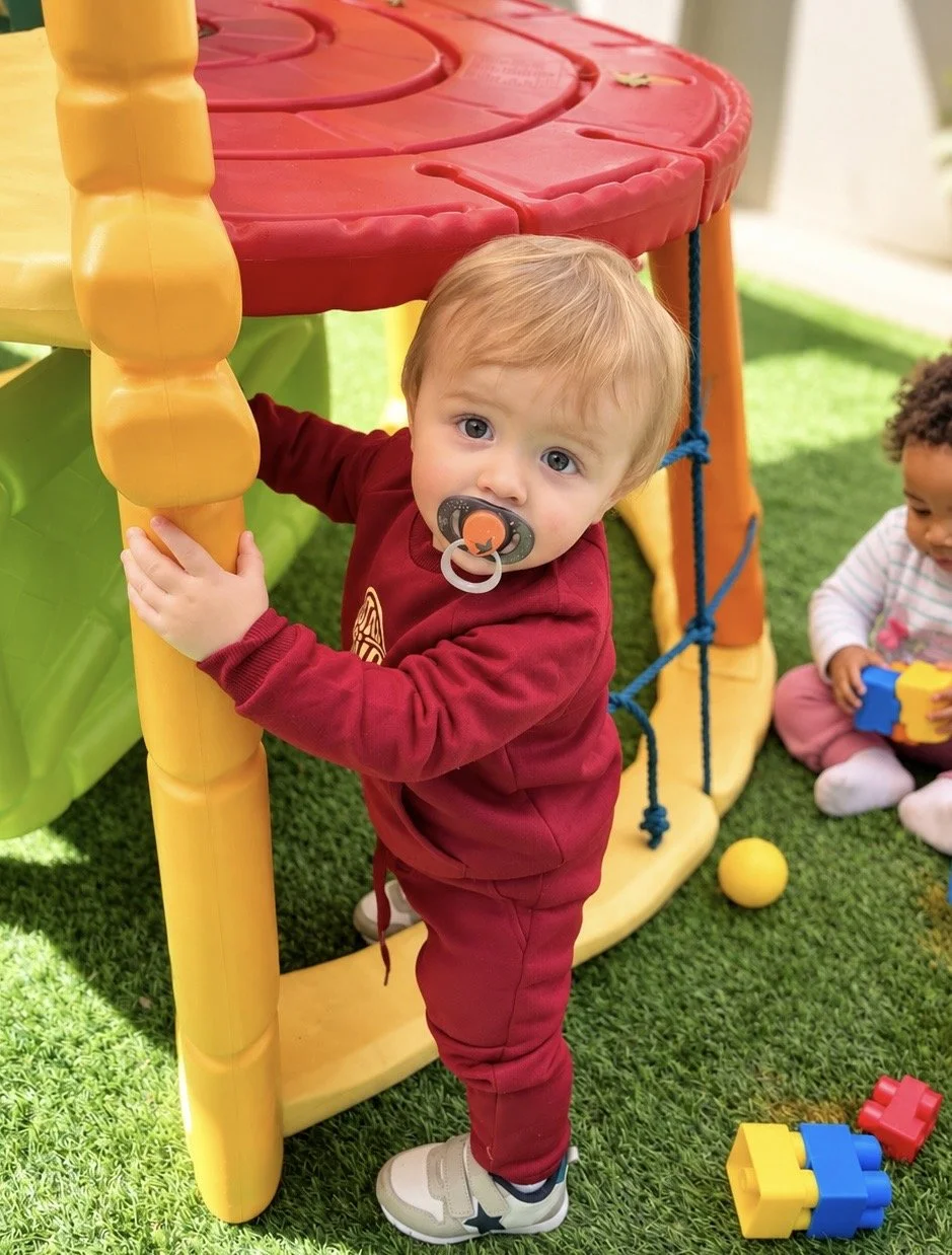 A young child with a pacifier in their mouth holding onto a yellow plastic support pole of a colorful outdoor play structure, with another child playing nearby on green turf grass.