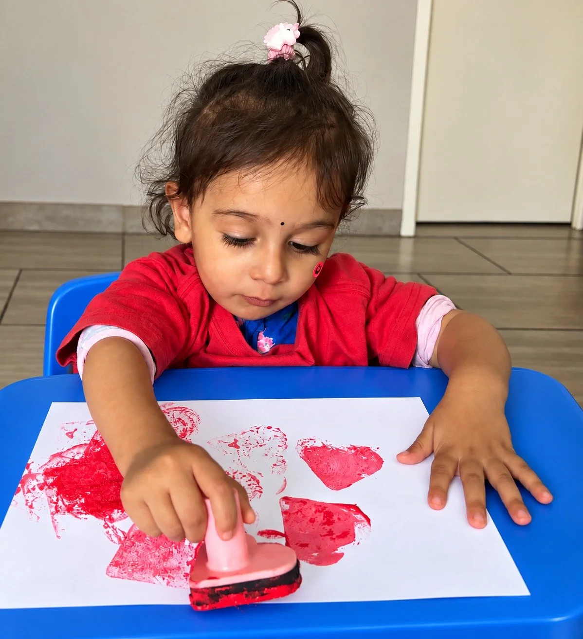 A young girl with dark hair in a ponytail with a pink hair tie stamping a red heart on a white sheet of paper, sitting at a blue table in a room with beige tiled flooring and a white door.