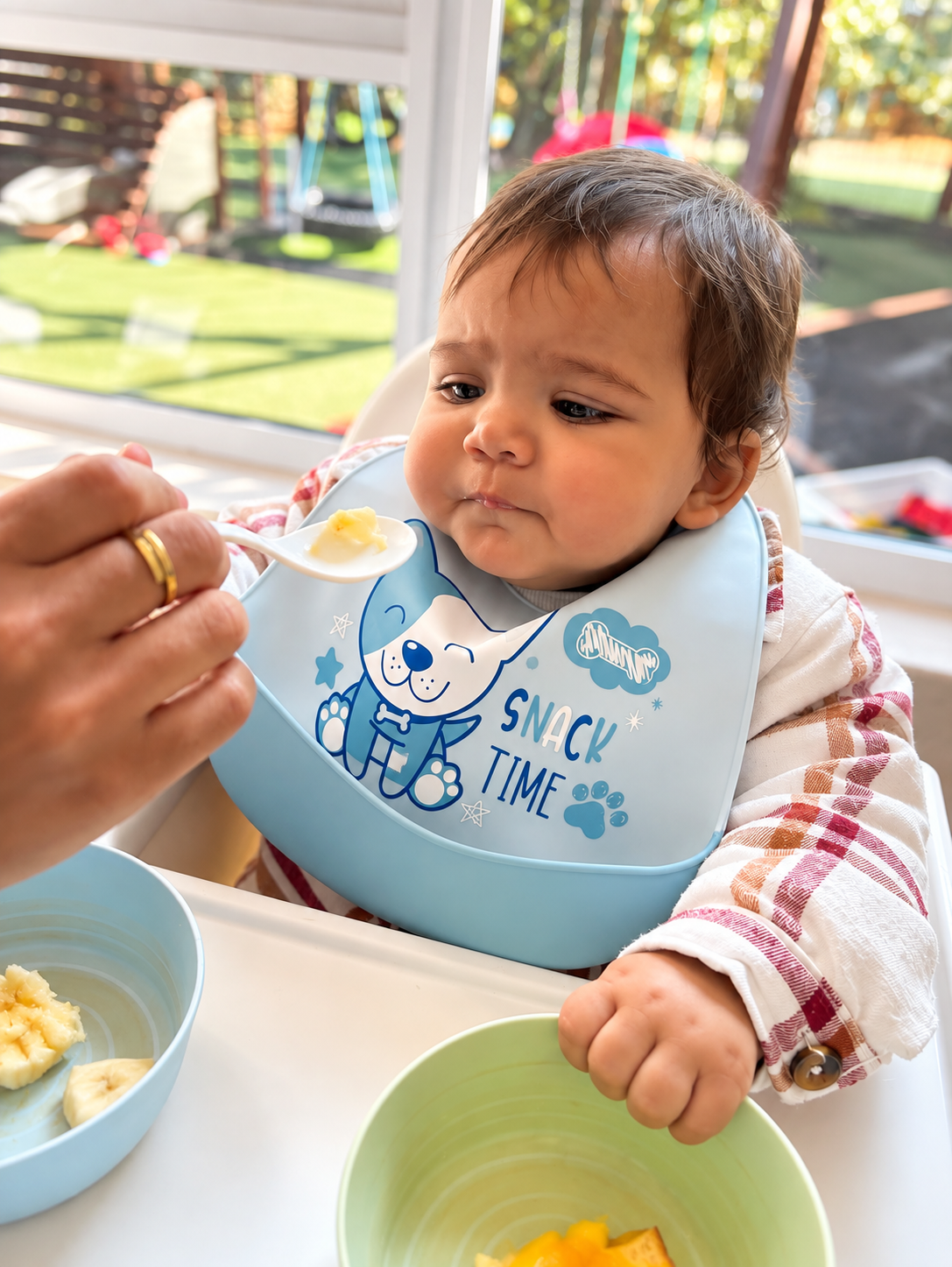 A young child sitting at a table during snack time, being fed banana on a spoon. The child wears a bib that says 'Snack Time' with a cartoon dog. There are bowls with food in front of the child, and a glass door with a view of a backyard with grass, 