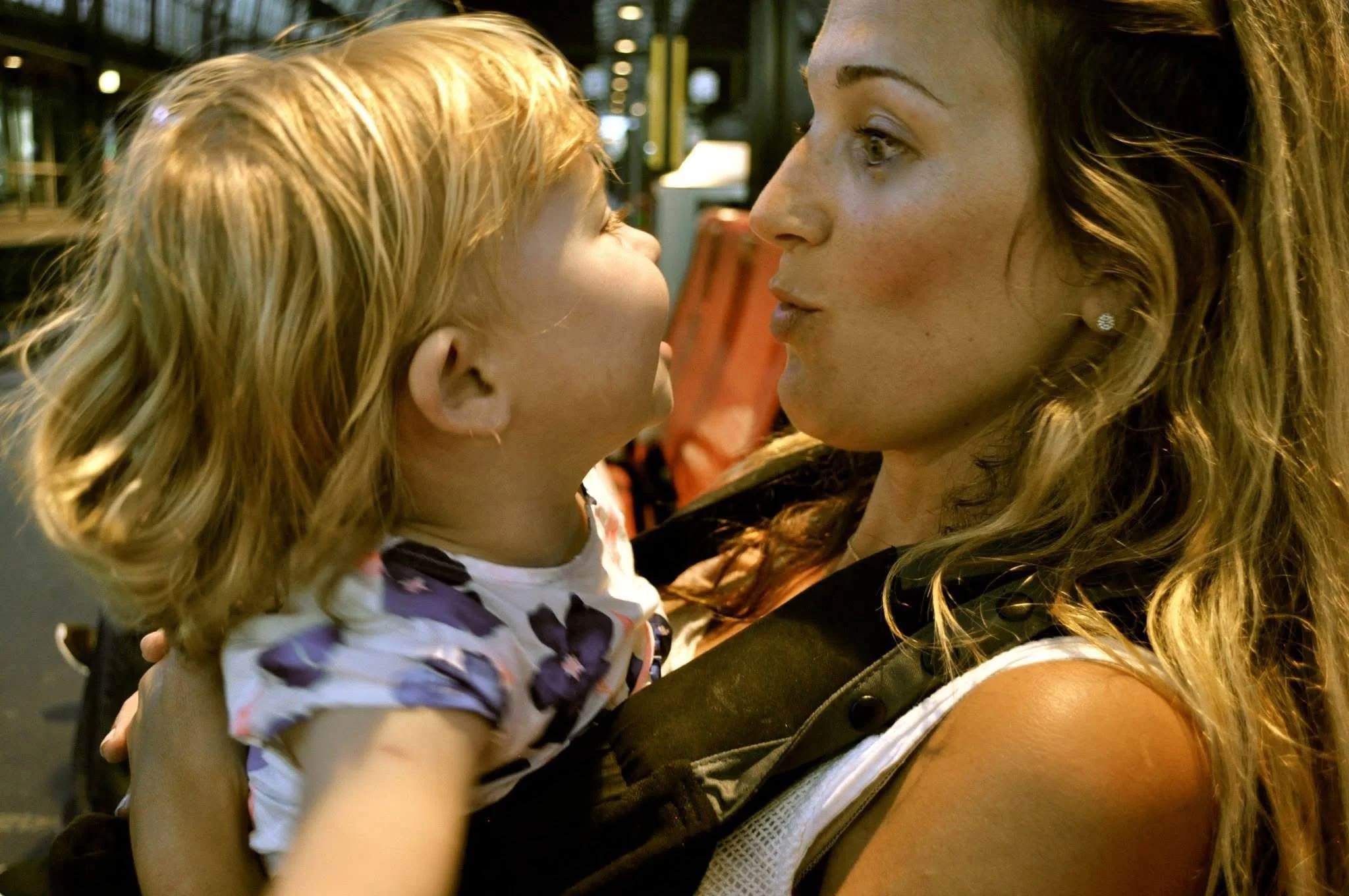 A woman with curly hair holding a young girl with blond hair close, both facing each other in an indoor setting.