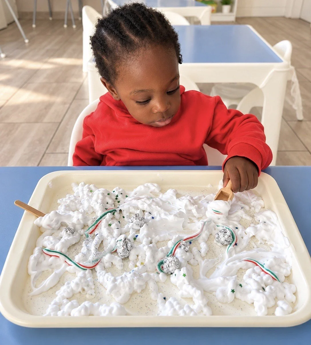 A young child in a red hoodie playing with a sensory tray filled with foam, silver balls, and craft decorations, using a small wooden tool, sitting at a blue table in a bright room.