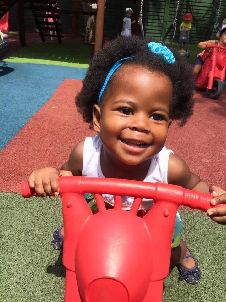 A young girl with curly hair and a blue headband smiling while holding onto a red play motorcycle at a playground. In the background, other children are playing, some on swings and others riding tricycles.