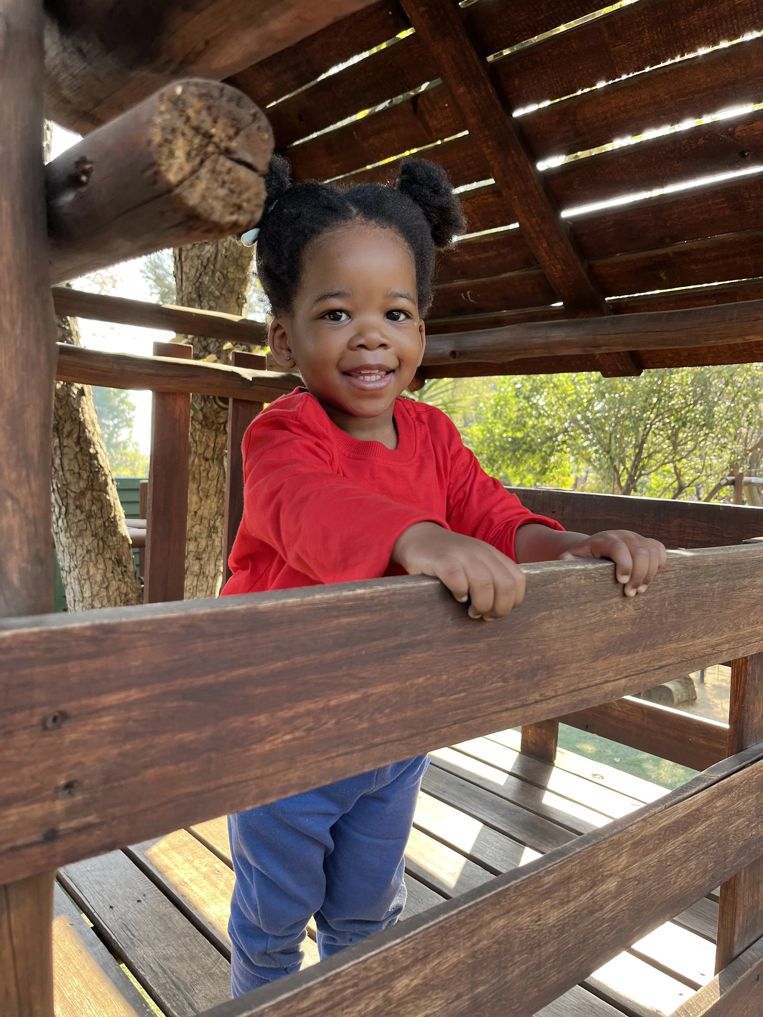 A young girl with dark hair styled in puffs, wearing a red long-sleeve shirt, is smiling and leaning on a wooden railing of a treehouse, with trees and greenery in the background.