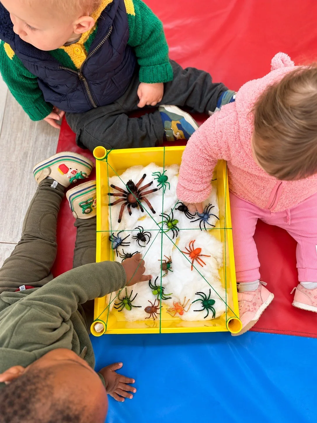 Children playing with plastic toy spiders and bugs in a yellow tray filled with cotton, with green string around the tray, on a padded surface.
