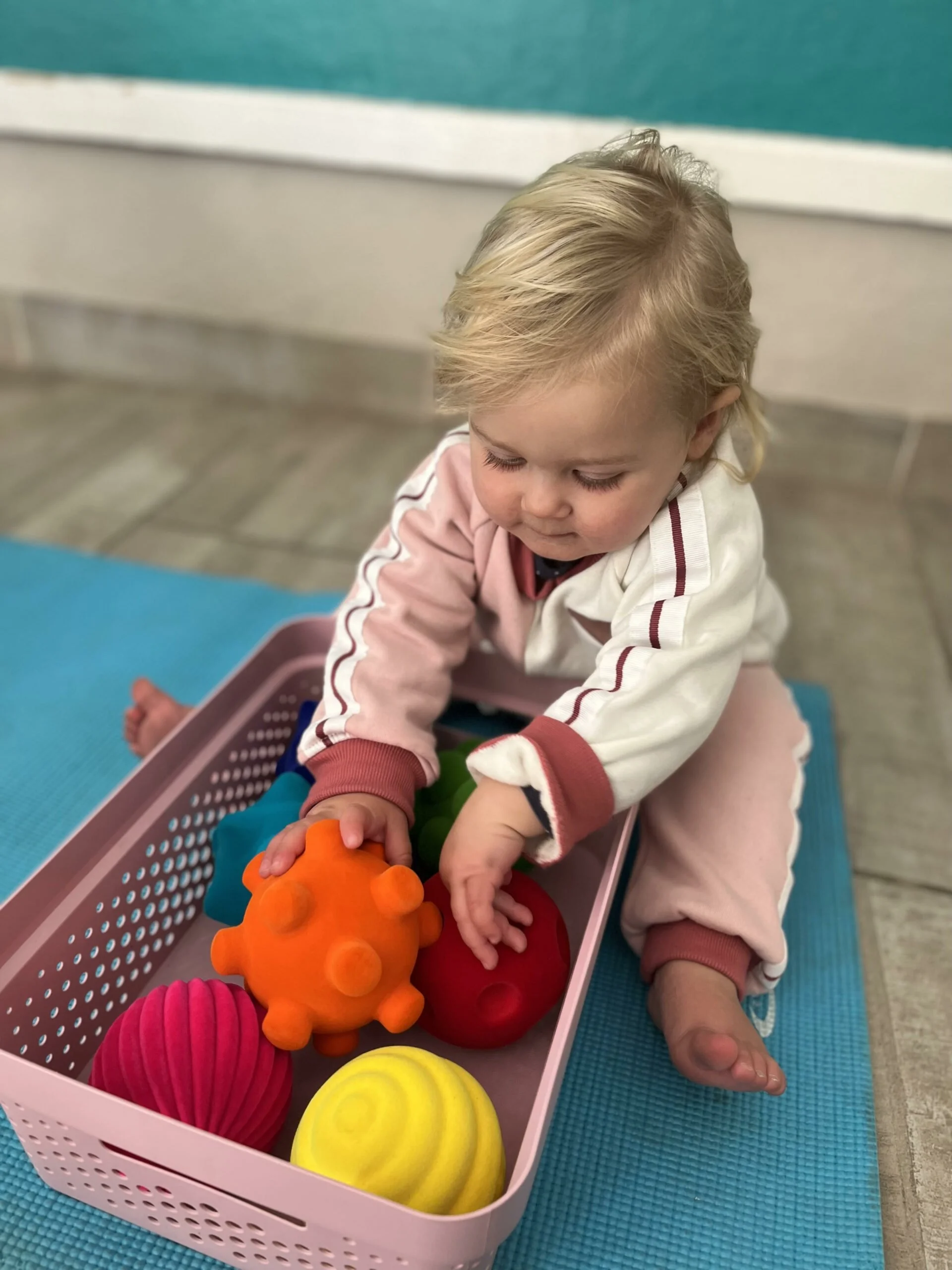 Young child with blond hair playing with colorful toys in a pink basket on a blue mat.