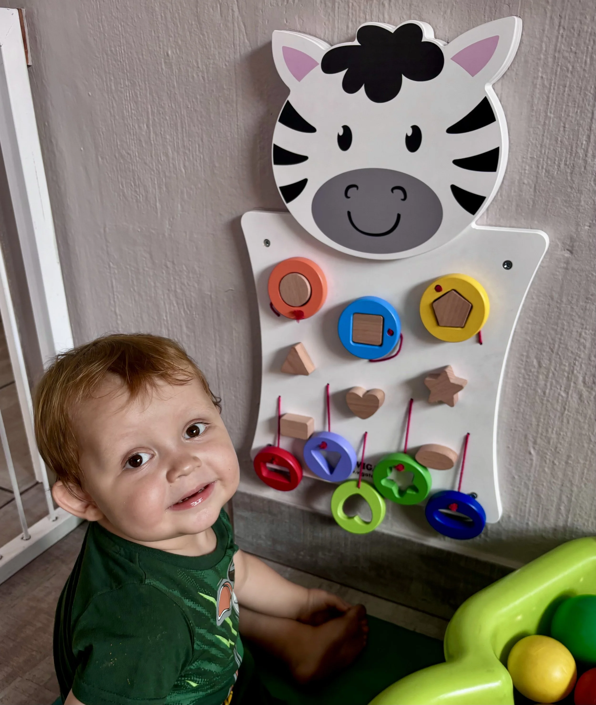 A young boy with brown hair and a green shirt smiling at the camera, seated next to a wall-mounted, zebra-shaped wooden activity board with colorful circular and geometric wooden pieces.