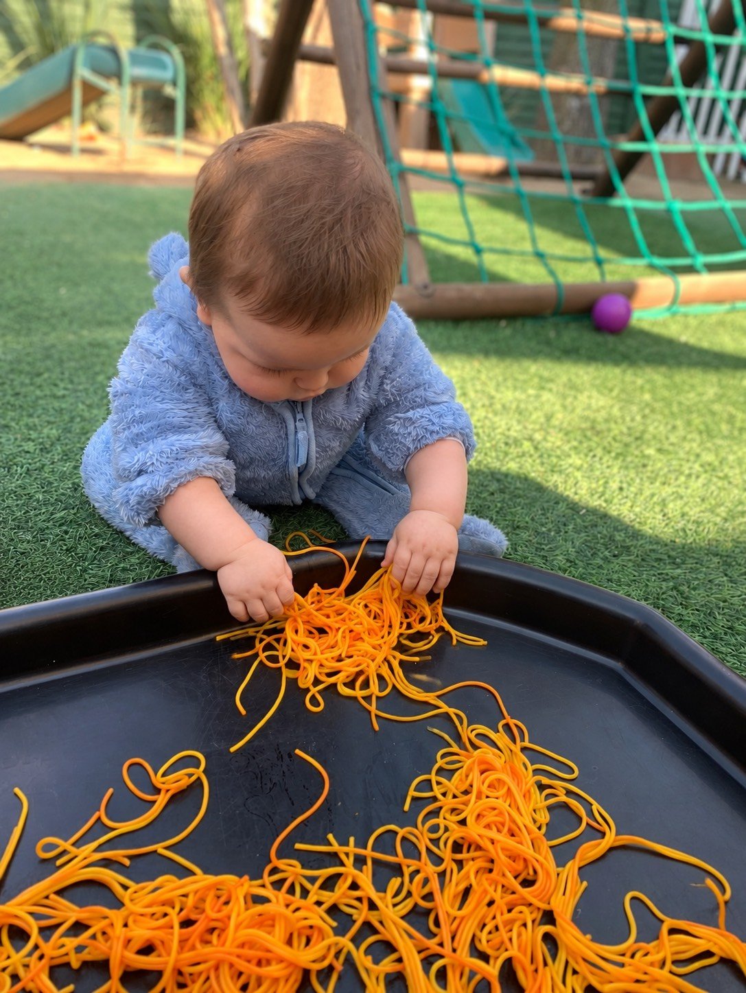 A young child in a blue, fuzzy jacket playing with orange rubber bands or noodles on a black tray outdoors, with a playground and green turf visible in the background.