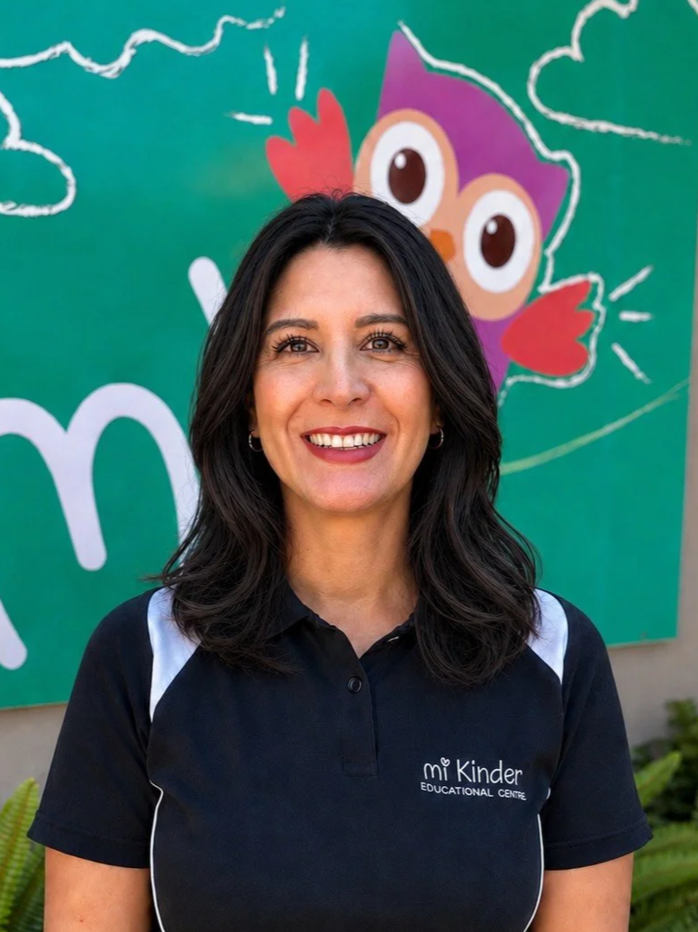 A smiling woman with dark hair and hoop earrings standing outdoors in front of a colorful cartoon owl mural. She is wearing a black polo shirt with a logo that reads 'Mi Kinder Educational Centre'.