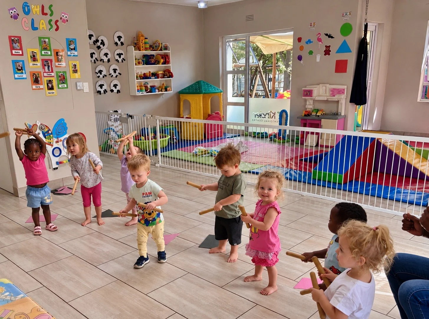 Children participating in a classroom activity with wooden sticks, standing on pink mats in a colorful indoor playroom, with toys and play structures in the background.